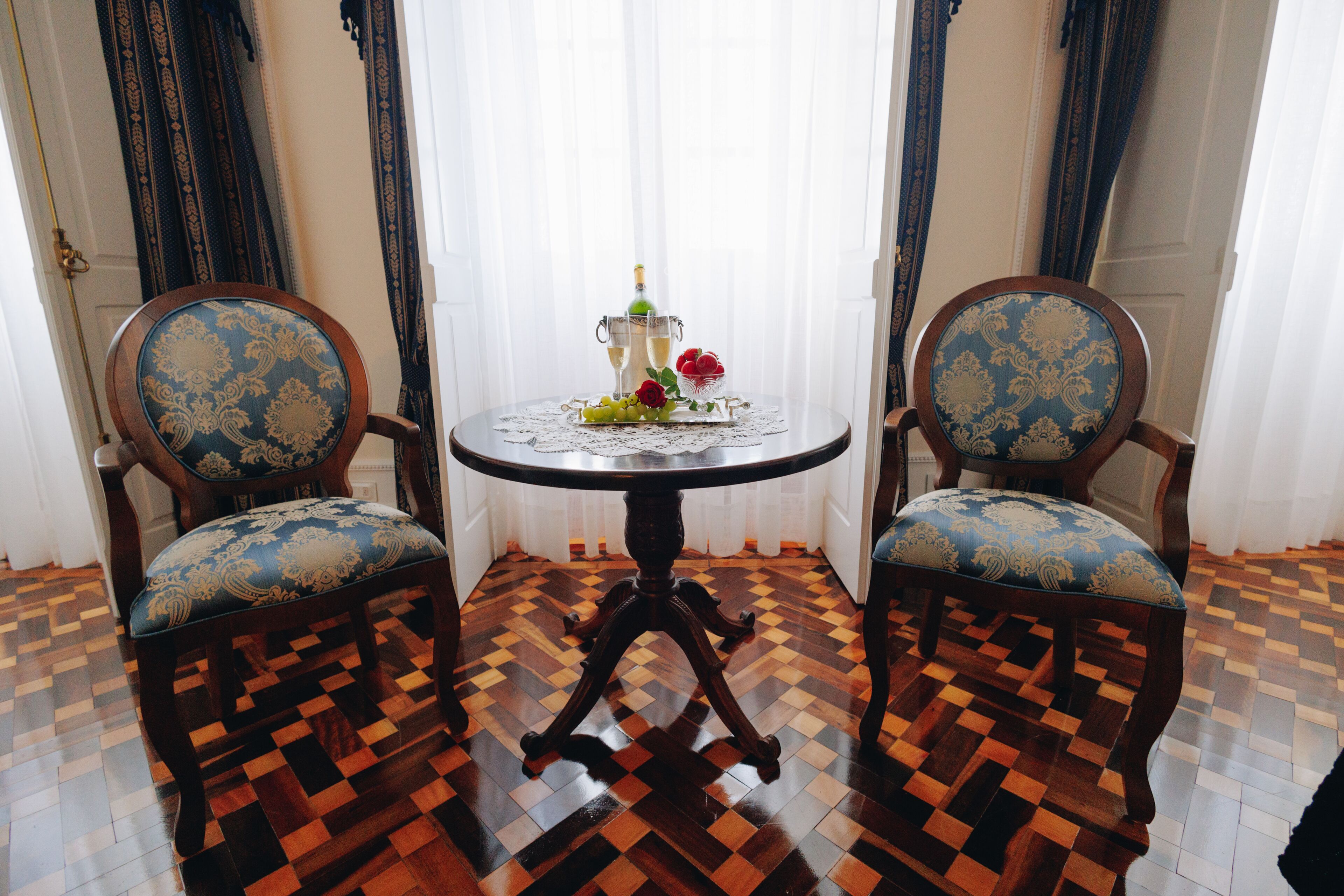 Una habitación con piso de madera a cuadros, dos sillones tapizados con estampado azul y dorado, una mesa redonda con un posavasos de encaje y una botella de champán.