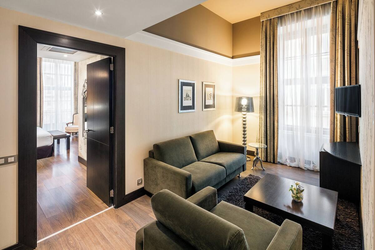A modern hotel room with a sofa, armchairs, a coffee table, and a TV.