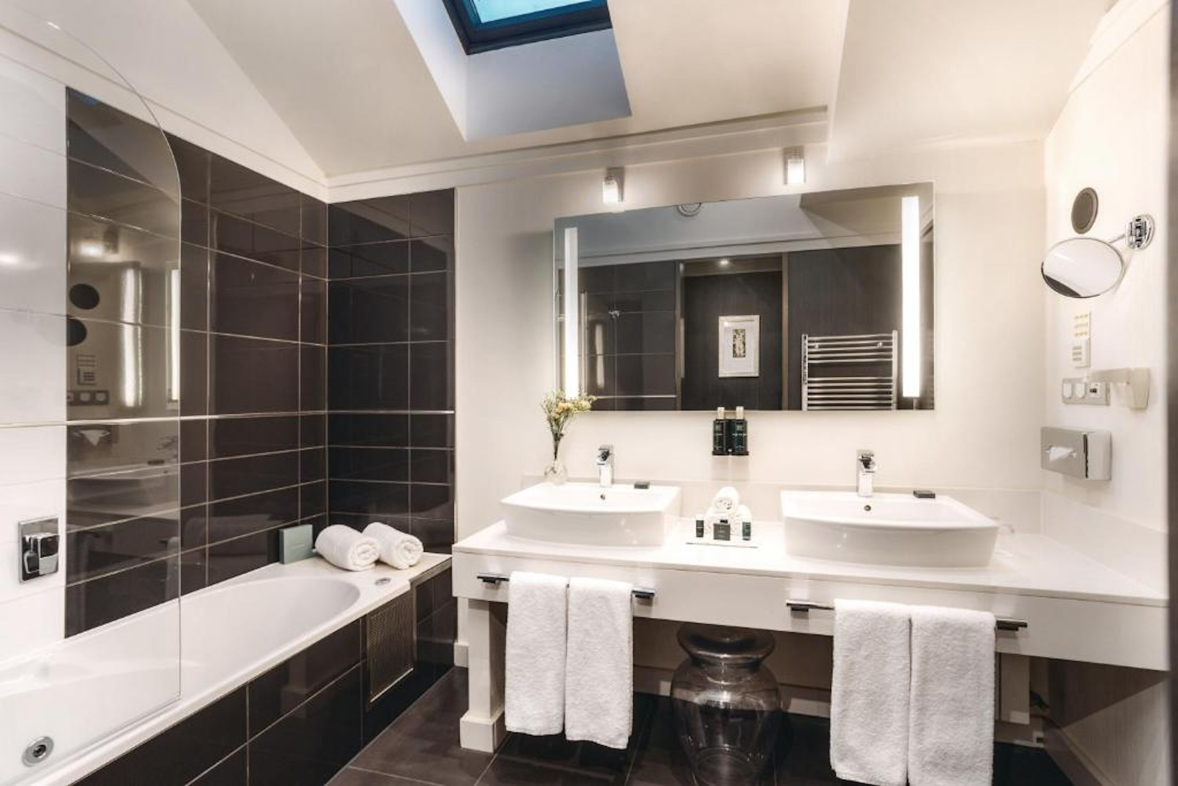 Un baño moderno con tragaluz, dos lavamanos, un espejo grande y una bañera.
