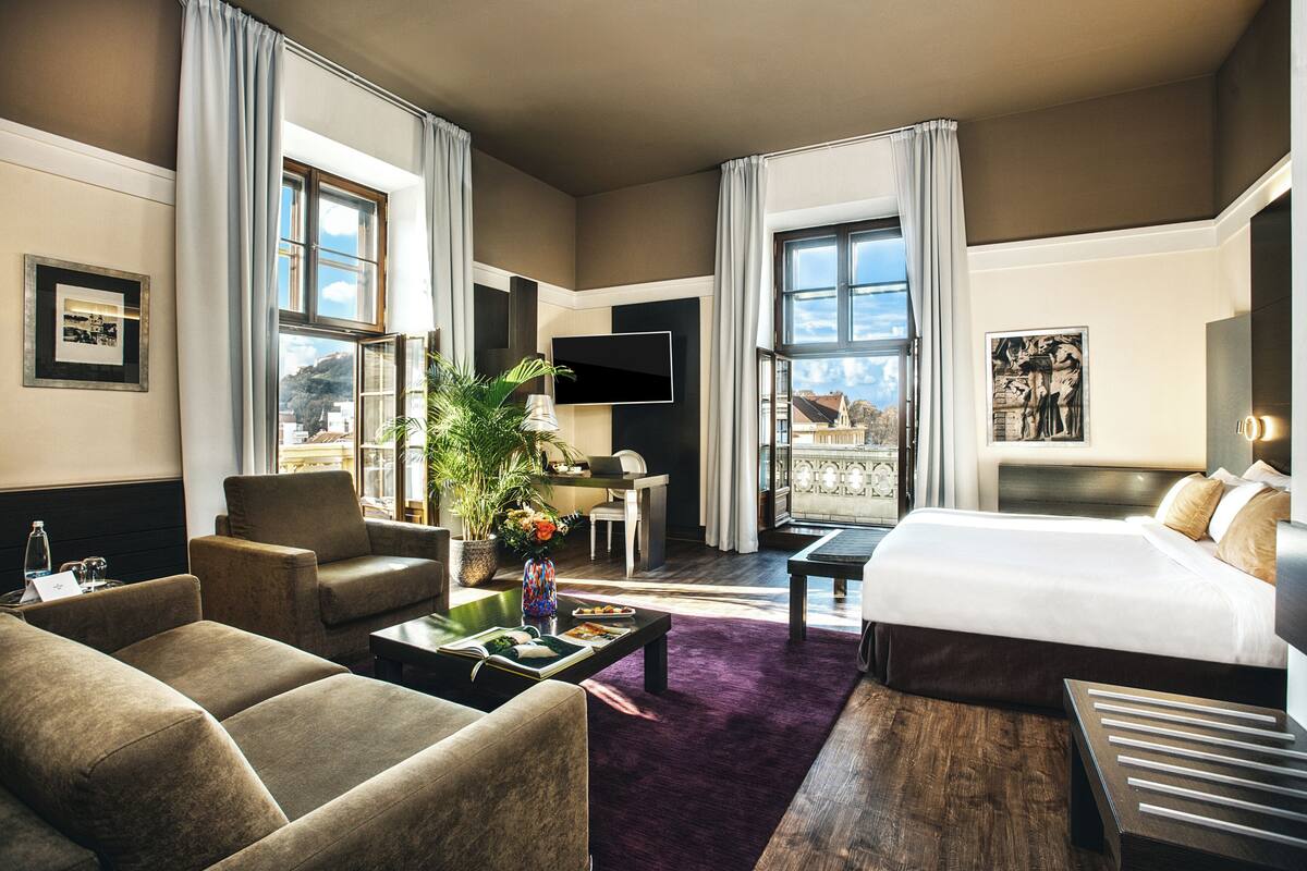 A hotel room with a large bed, a sofa, a coffee table, and a desk. There is a TV mounted on the wall, a plant on the desk, and a window with curtains.