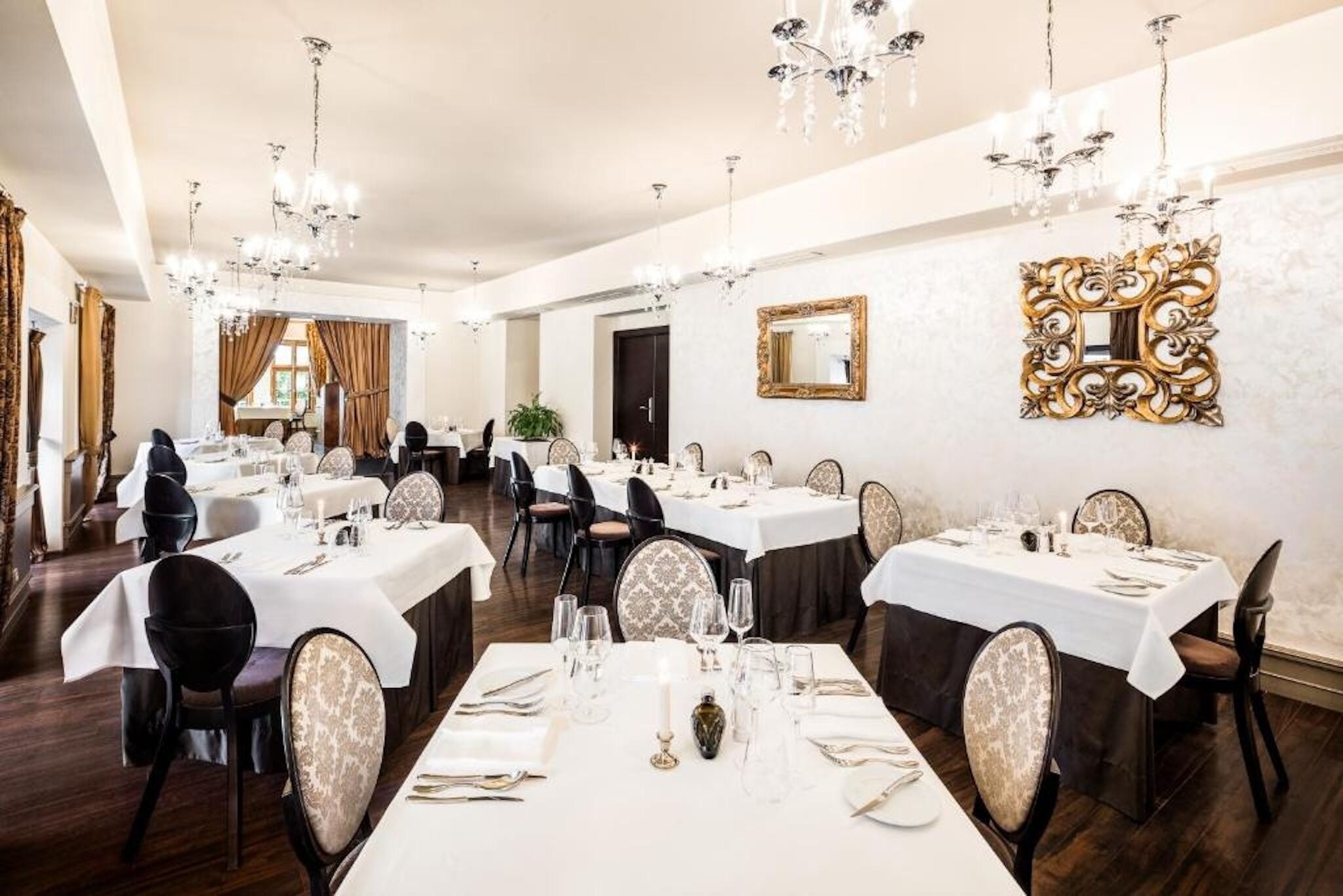 A formal dining room with multiple tables set for service, elegant chandeliers, and ornate wall mirrors.