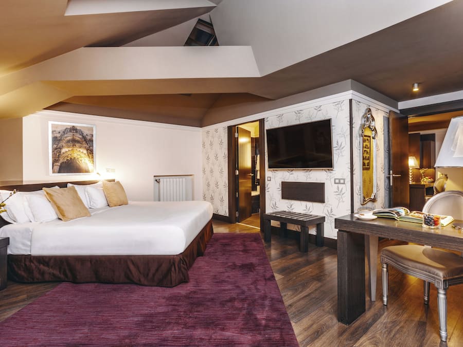 A modern hotel room with a large bed, a desk, a television, and a fireplace.