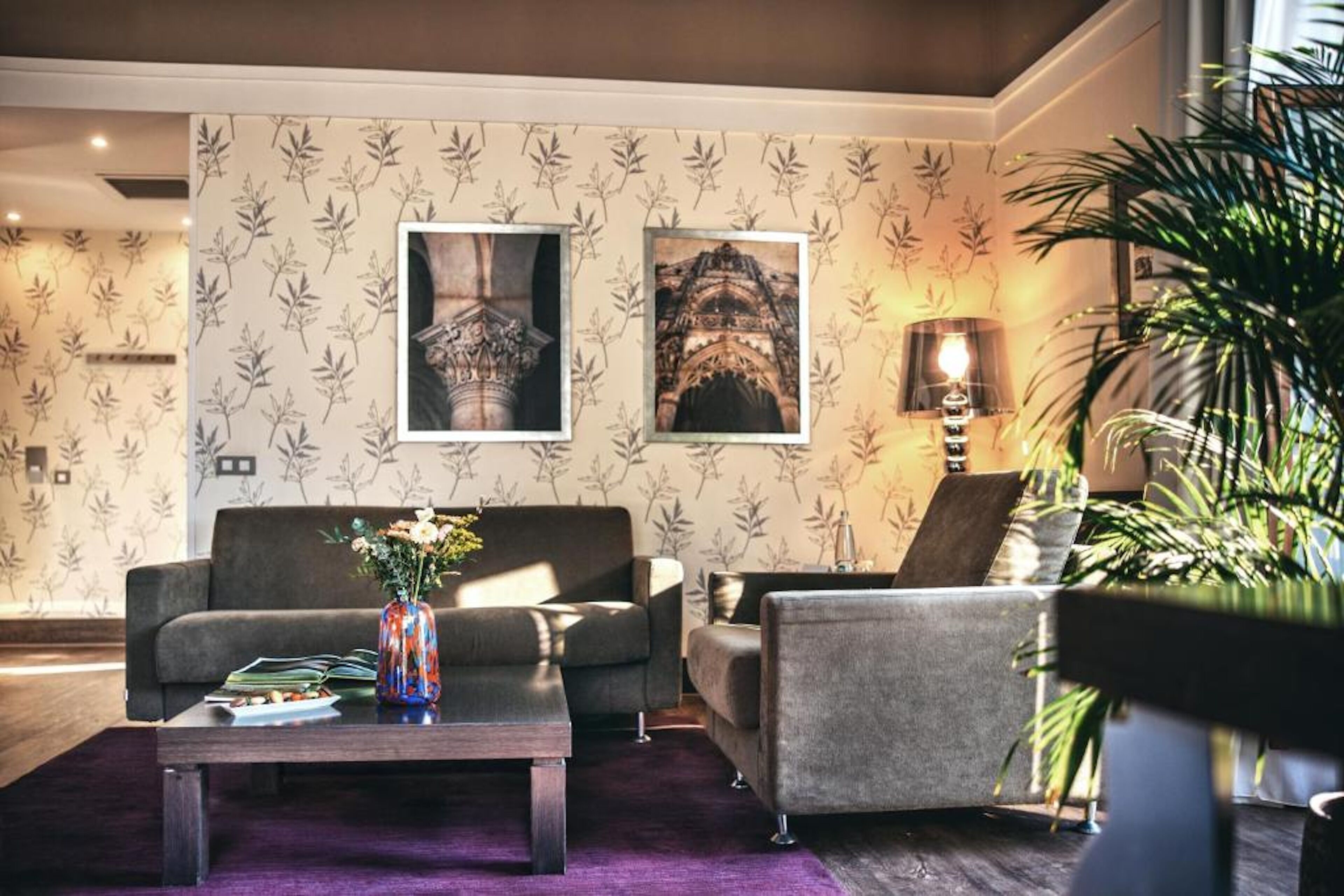 A modern living room with a sofa, armchairs, a coffee table, and a vase with flowers. There are framed pictures on the wall and a lamp on a side table.