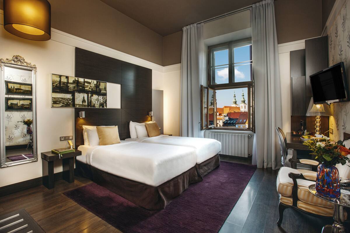 A hotel room with a large bed, a desk with a TV, a window with a view of buildings, and a mirror with pictures on the wall.