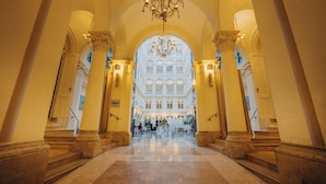 A grand hallway with a chandelier, columns, and a view of a building with multiple windows. - Grand Palace Brno - Small Luxury Hotels of the World (Brno)