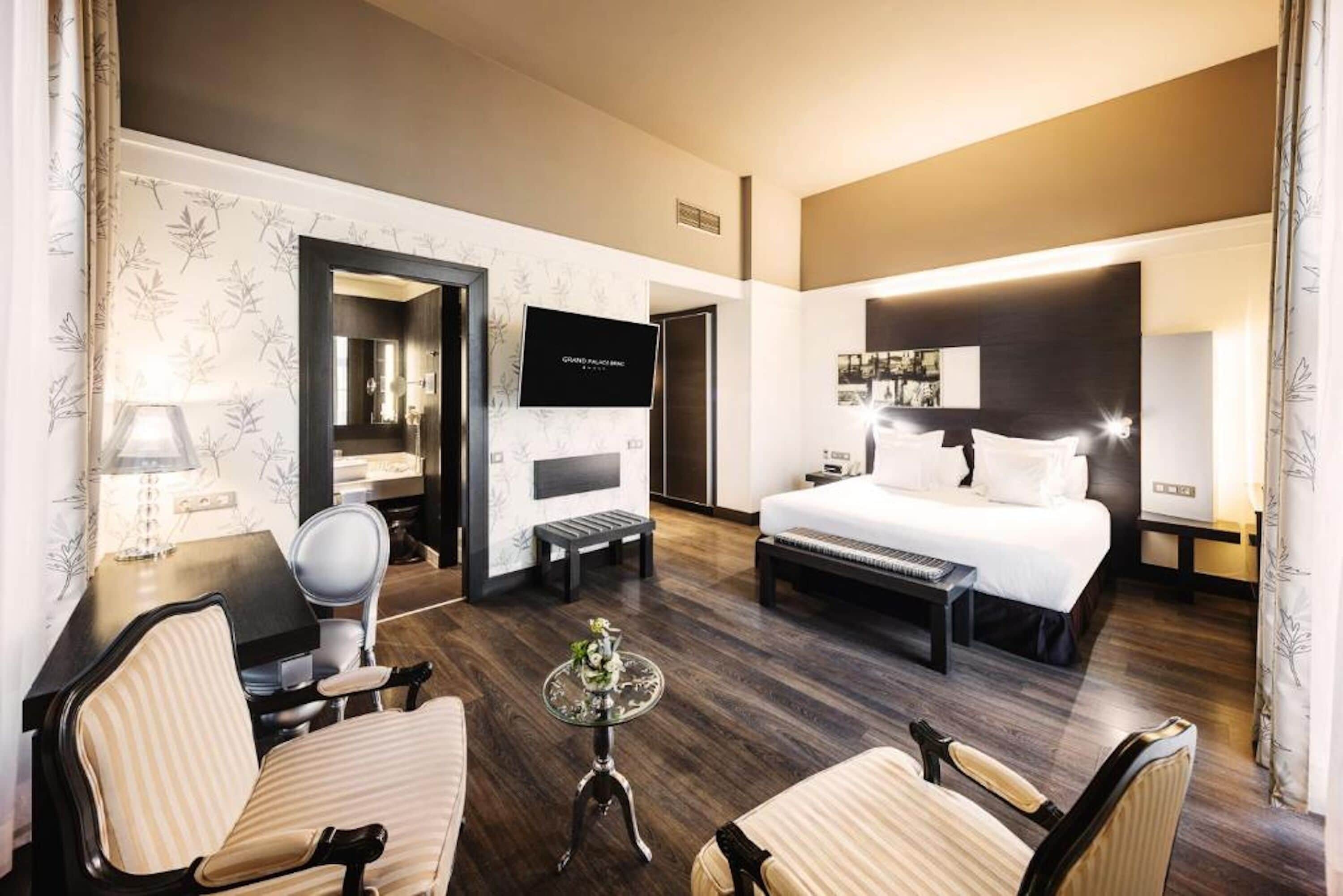 A hotel room with a bed, two armchairs, a small table, a TV, and a mirror.