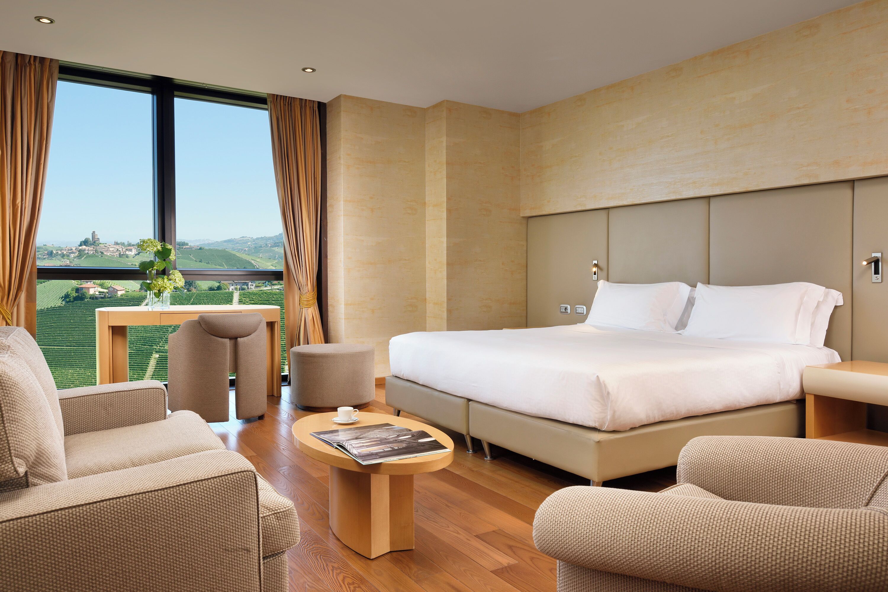 A modern hotel room with a large bed, a sofa, a small table, and a view of a green landscape through large windows.