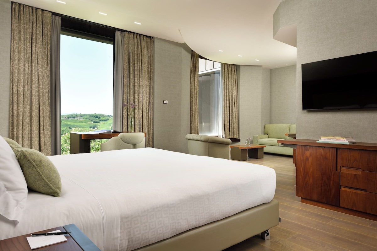 A modern hotel room with a large bed, a desk, a sofa, and a TV mounted on the wall.