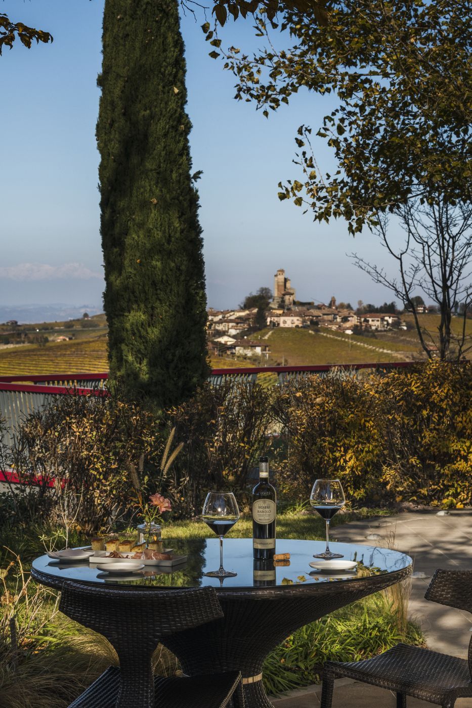 Mesa redonda com uma garrafa e copos de vinho, disposta ao ar livre com vista para uma aldeia e uma torre ao fundo.