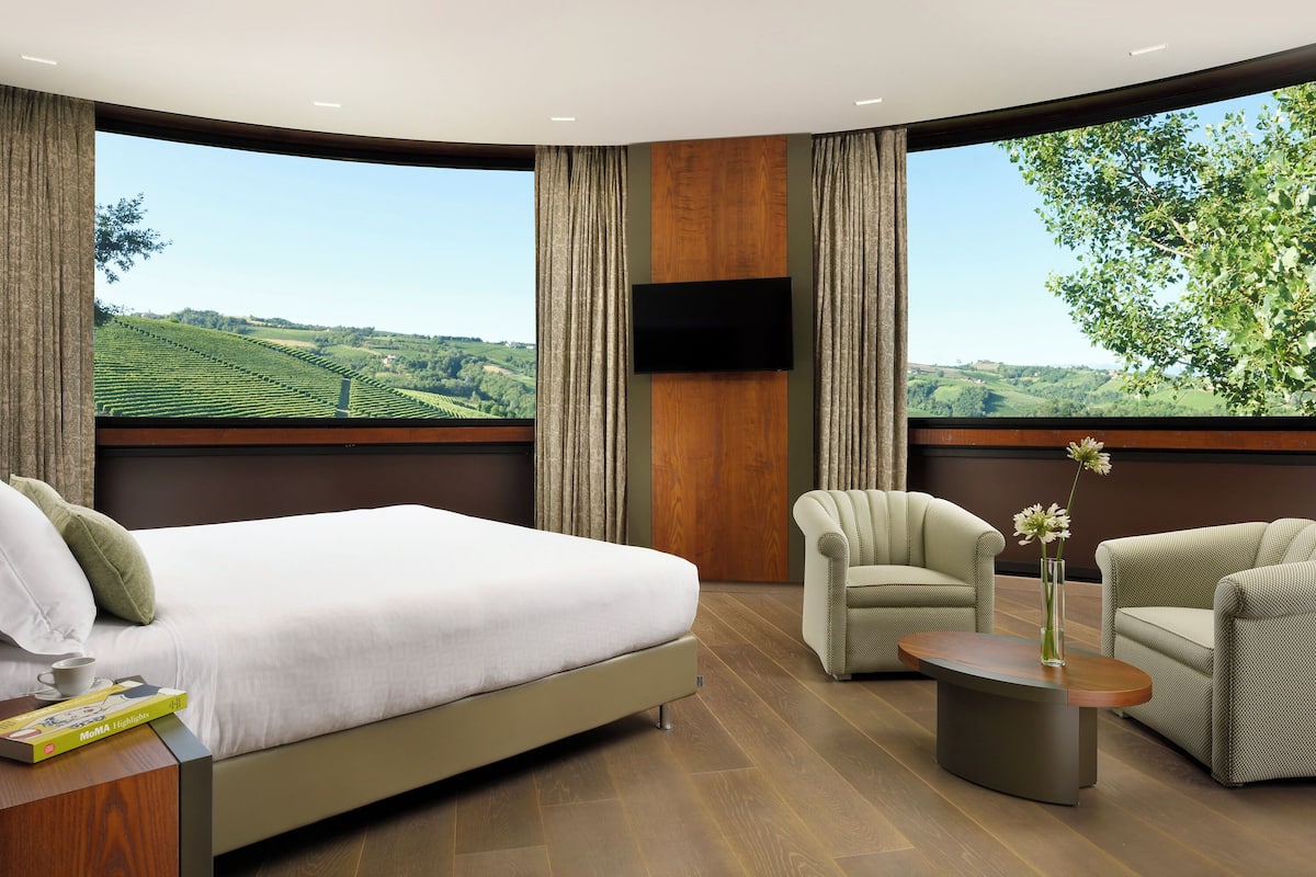 A modern hotel room with a large bed, two armchairs, a coffee table, and a TV mounted on the wall.