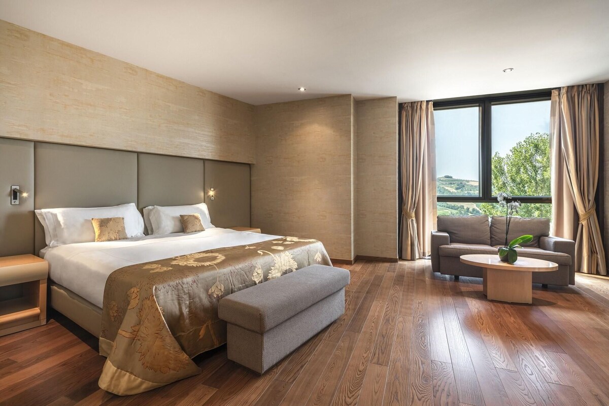 A modern hotel room with a large bed, a sofa, a coffee table, and a view of the outdoors.