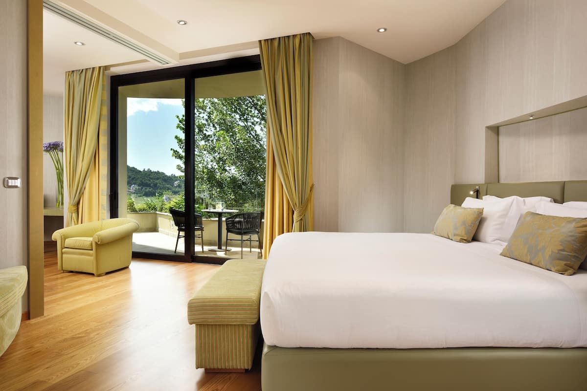 A modern hotel room with a large bed, a sofa, a dining area with a table and chairs, and a view of greenery outside.
