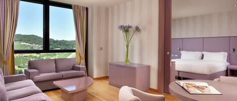 A modern hotel room with a large window, a sofa set, a small table, and a bed with a headboard.
