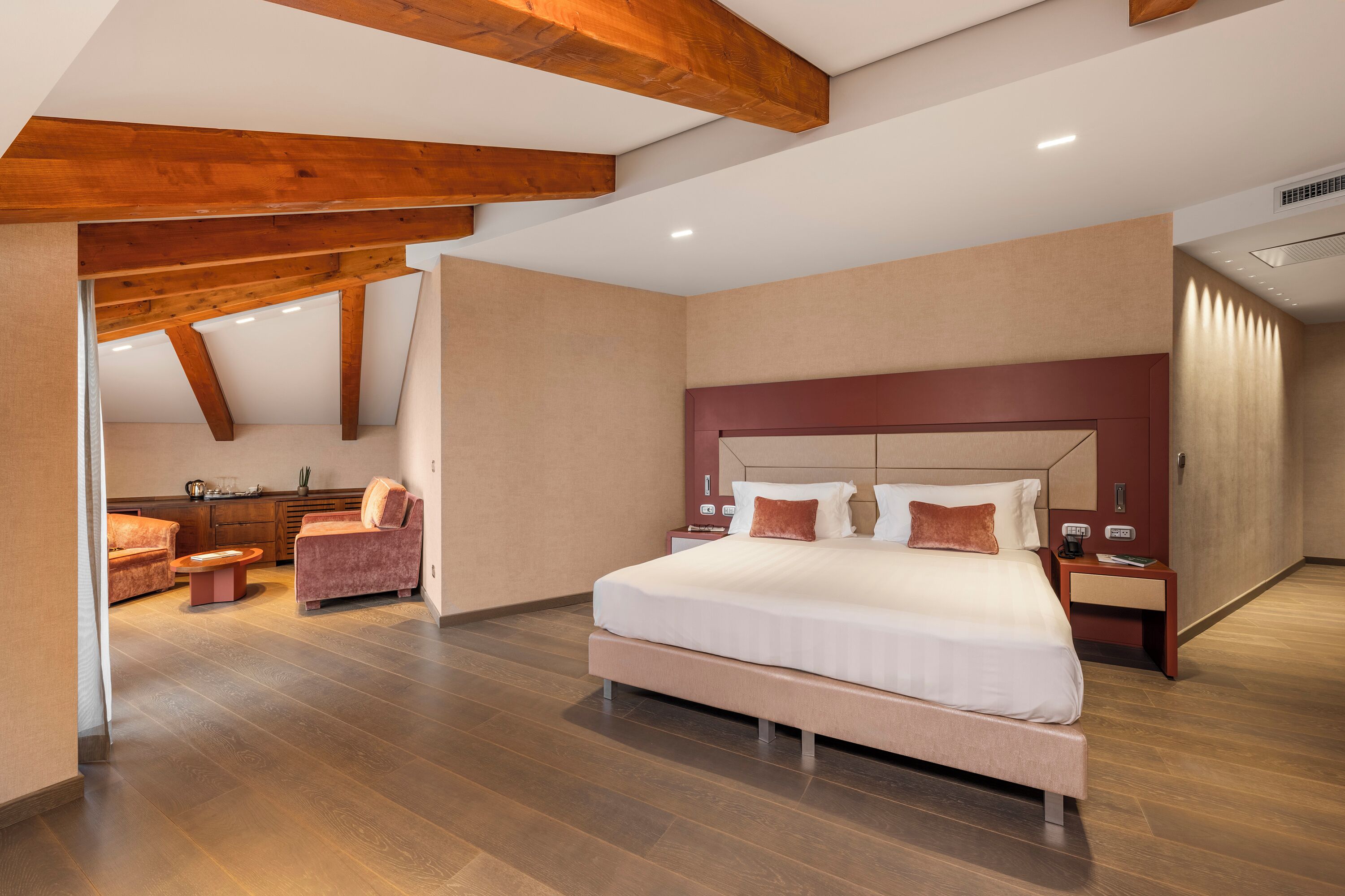 Ein modernes Hotelzimmer mit einem großen Bett, Holzbalken, einer Sitzecke mit Sofa und Sesseln sowie einem kleinen Tisch.