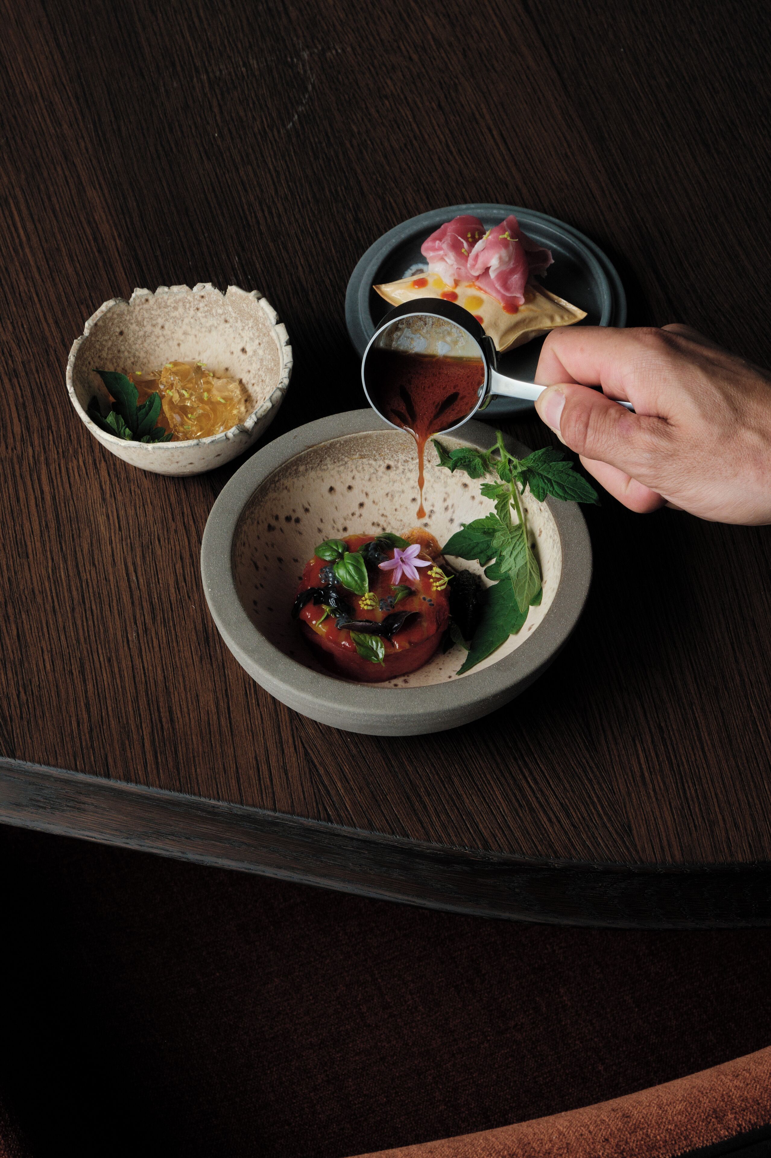 A hand pouring sauce into a bowl with a tomato and basil garnish.