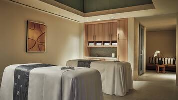 A hotel room with two beds, a small seating area, a bathroom with a shower, and a large window.