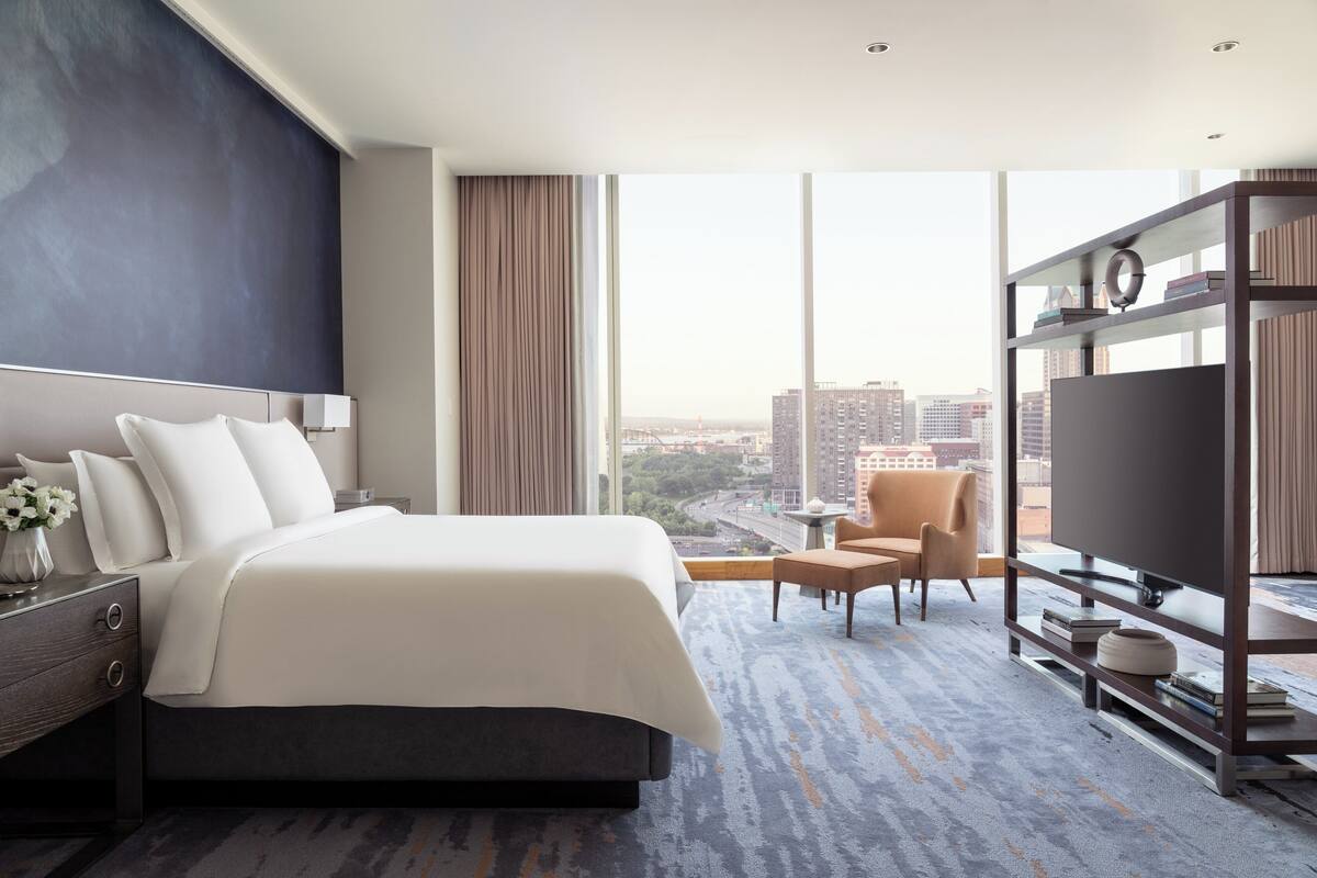 A modern hotel room with a large bed, a television, a bookshelf, and a city view.