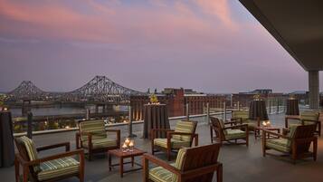 A rooftop terrace with wooden chairs and tables, a view of a bridge, and a cityscape in the background.