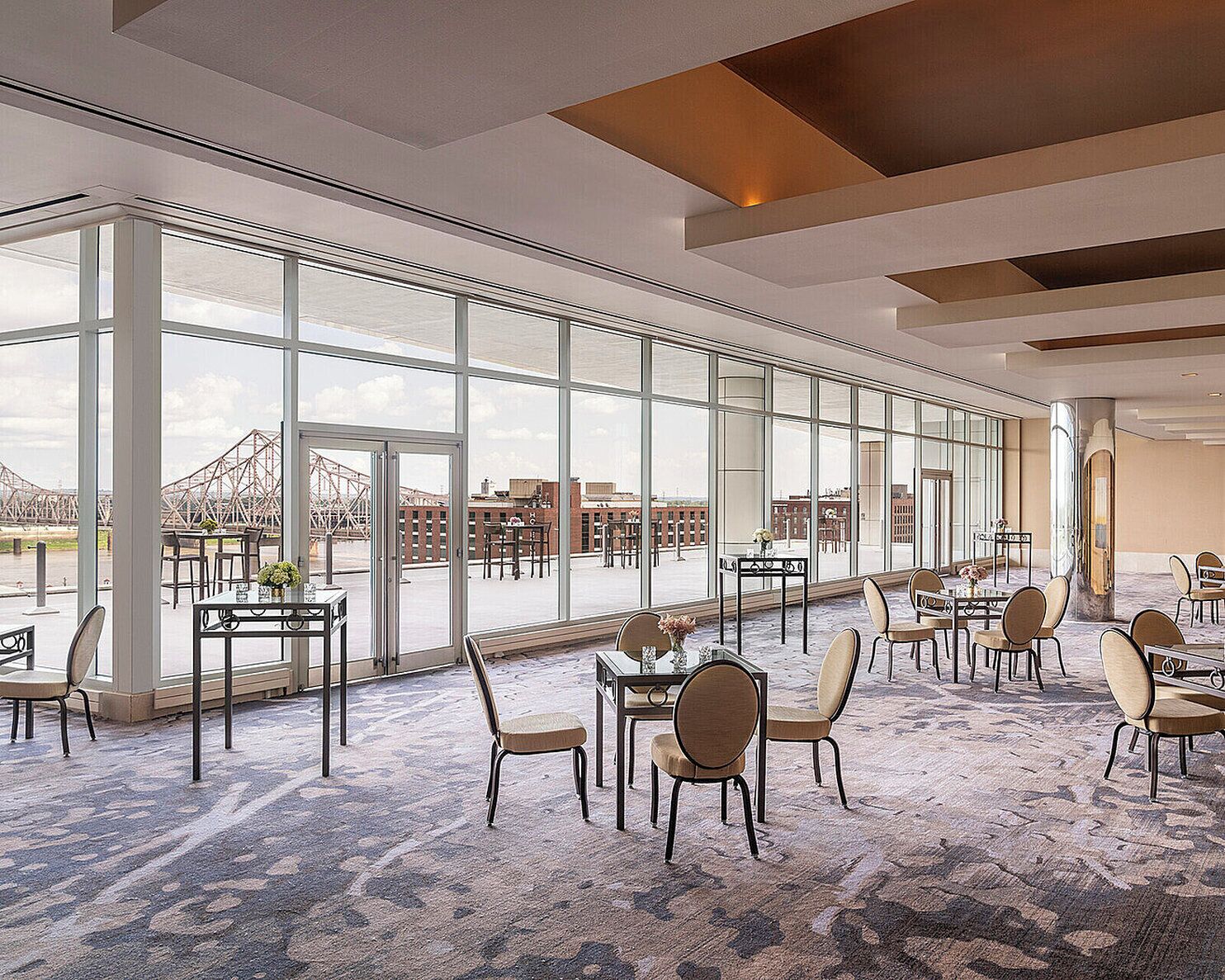 A spacious indoor area with large windows, multiple tables and chairs, and a view of a bridge and buildings outside.