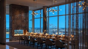 A modern dining area with a long table set for a meal, large windows offering a view of a bridge, and a stone accent wall.