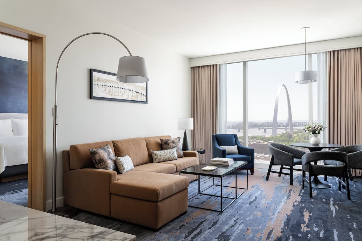 A modern hotel room with a sofa, armchair, and dining table. A large window offers a view of a bridge.