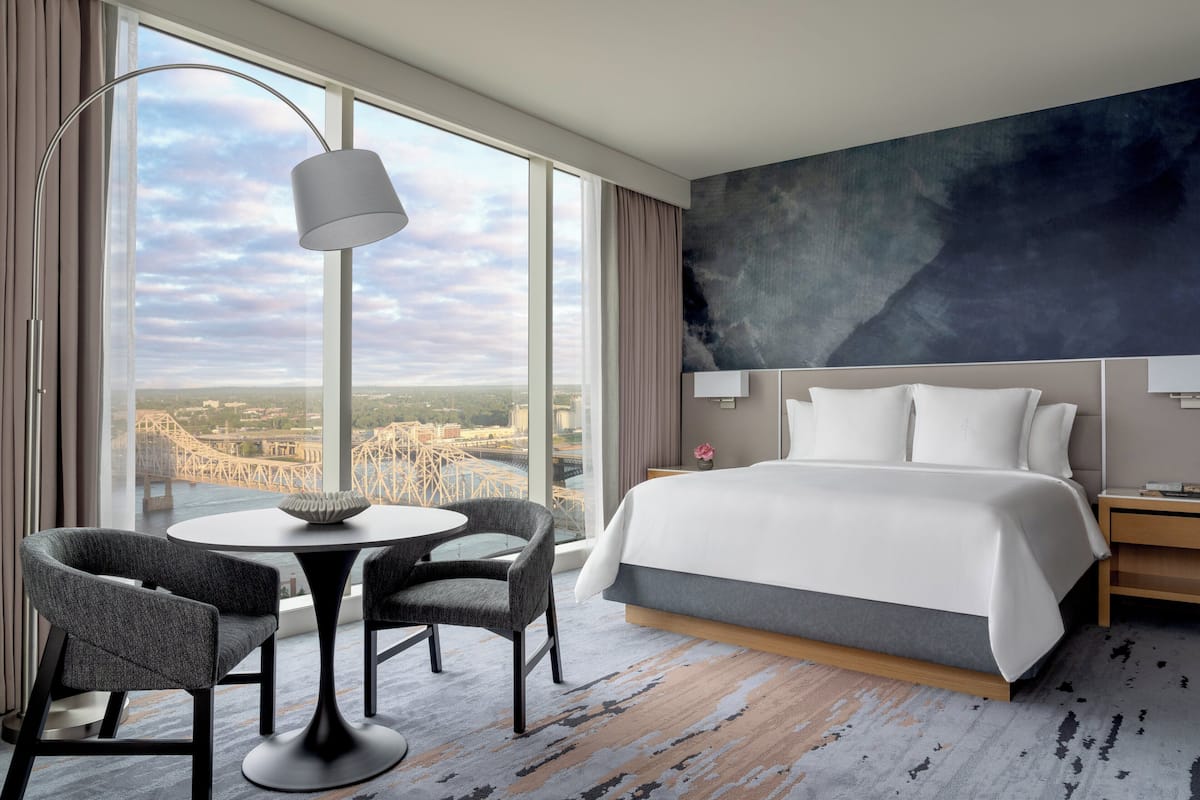 A modern hotel room with a large bed, a round table with two chairs, a view of a bridge, and a cityscape.