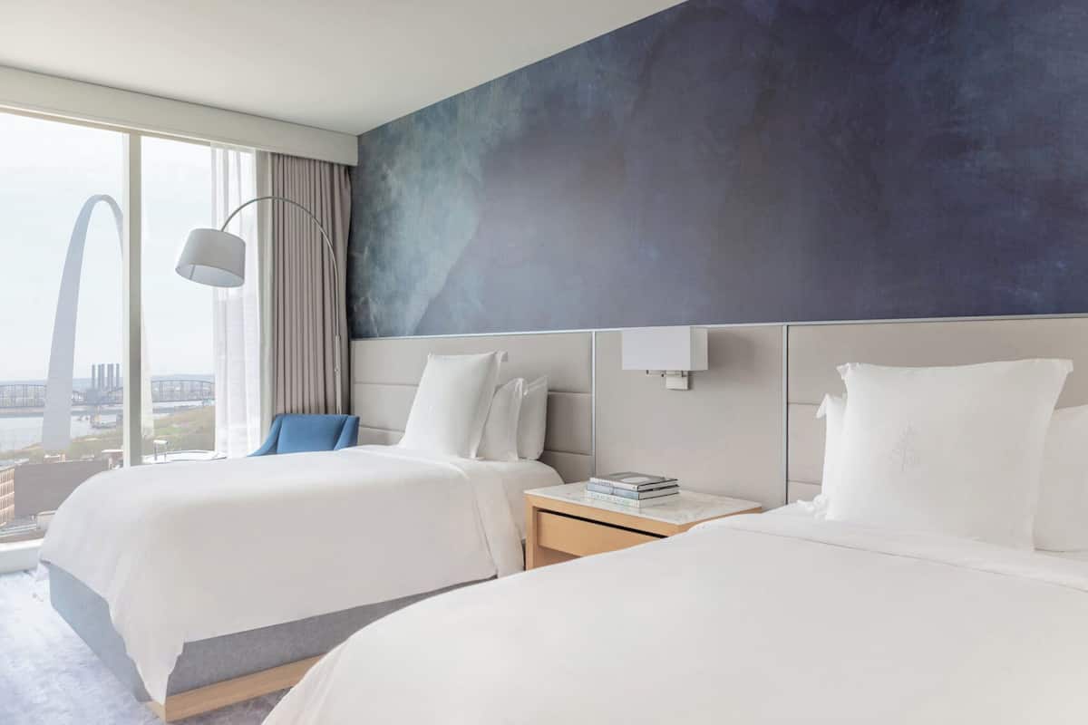 A hotel room with two beds, a view of a cityscape, a modern lamp, and a large window with curtains.