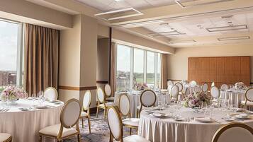 Un salón de banquetes con mesas redondas dispuestas para comer, ventanas amplias que ofrecen vista a la ciudad y un techo alto con iluminación empotrada.