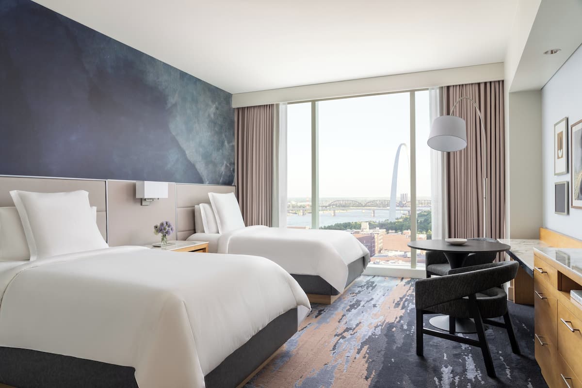 A hotel room with two beds, a dining table, a chair, and a view of a bridge.