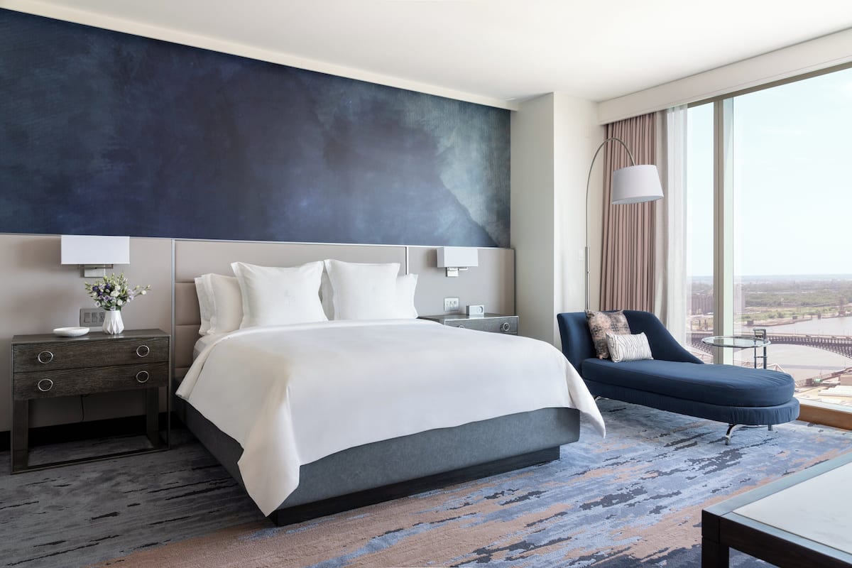 A modern hotel room with a large bed, a sofa, a nightstand with a lamp, and a view of the city through large windows.