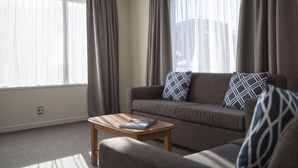 A modern living room with a grey sofa, two patterned cushions, a wooden coffee table, and sheer curtains on large windows. - Dunedin Motel and Villas (Dunedin)