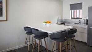 A modern kitchen with a white island, four bar stools, a bowl of fruit, and a window with a view of a brick building. - Dunedin Motel and Villas (Dunedin)