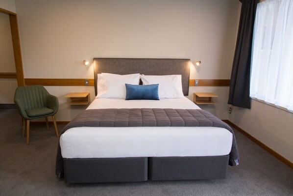 A large bed with a headboard, two bedside tables, a green chair, and a window with curtains. - Dunedin Motel and Villas (Dunedin)