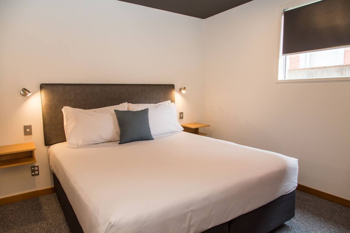 A neatly made bed with a dark headboard and white bedding, two bedside tables with lamps, and a window with a blind.