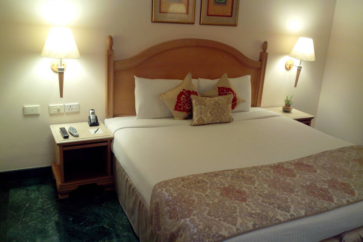 A hotel room with a neatly made bed, two bedside lamps, a nightstand with a phone and remote controls, and a small plant on the nightstand.