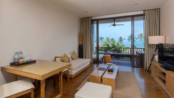 Quarto de hotel moderno com piso de madeira, sofá, mesa de jantar, televisão e varanda com vista para o oceano e palmeiras.