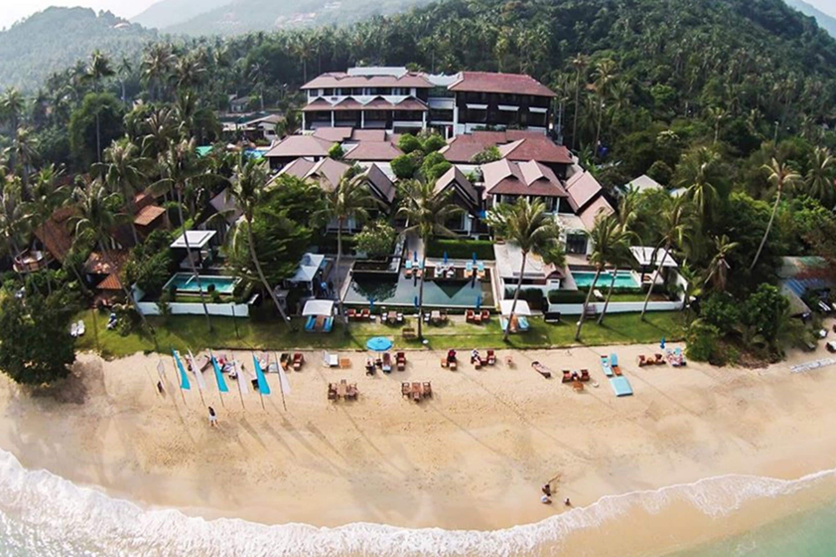 Vista aérea de un resort con varias construcciones, una playa y palmeras.