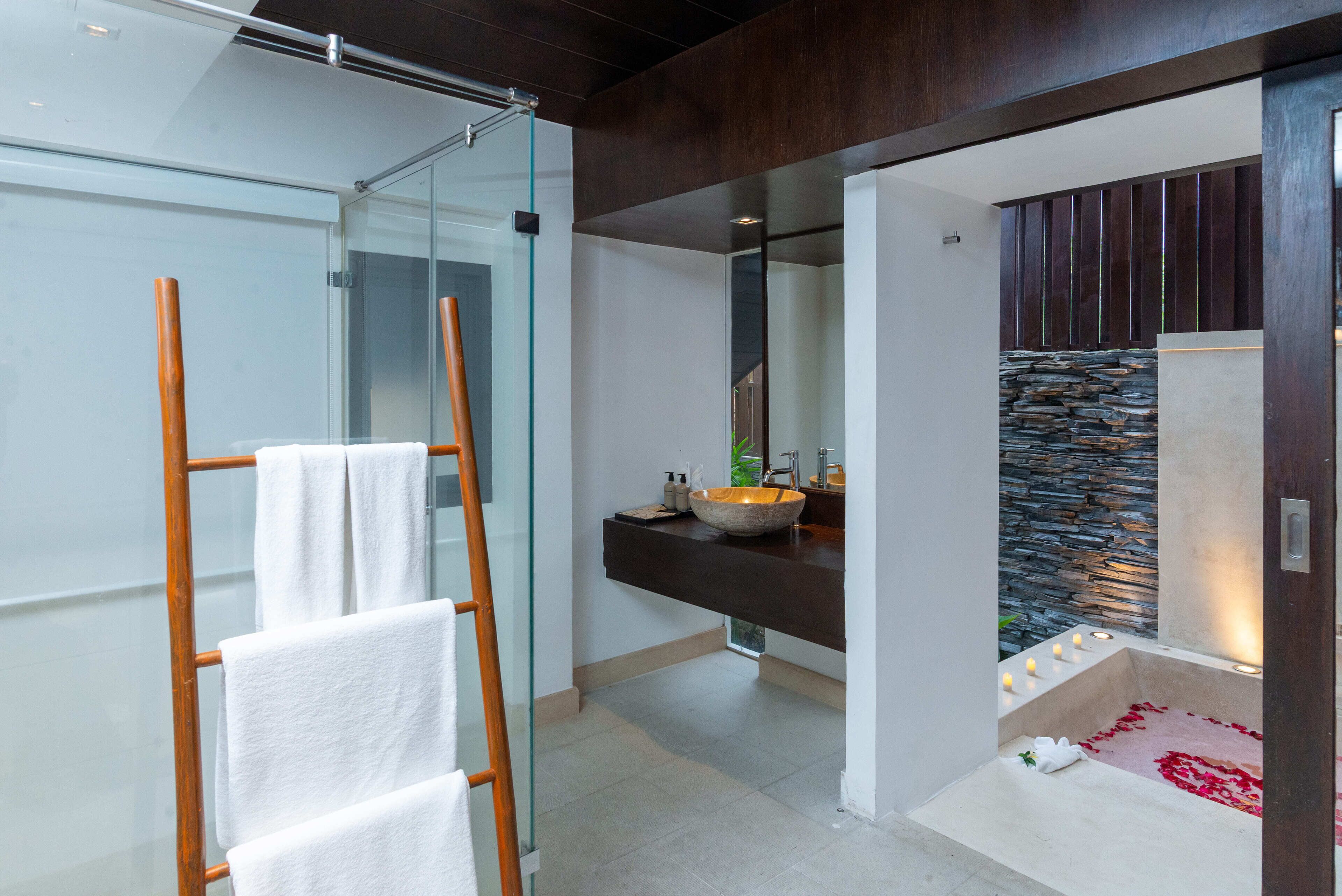 Un baño moderno con cabina de ducha de vidrio, toallero de madera, pared de acento de piedra y bañera con velas.