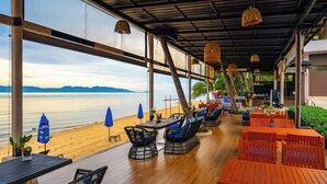 A beachside restaurant with outdoor seating, large windows overlooking the sea, and a covered area with hanging lights. - The Sea Koh Samui Resort & Residences by Tolani (Koh Samui)