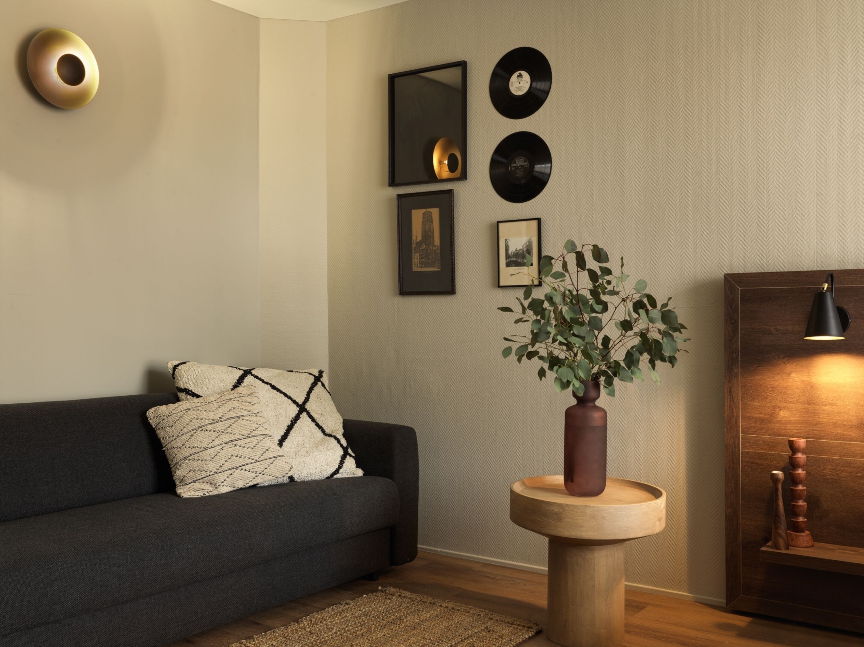 A modern living room with a dark grey sofa, a round wooden side table with a plant, and a wall decorated with framed pictures and vinyl records.