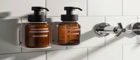Dos botellas de loción para manos de color ámbar con dosificadores negros, fijadas a una pared de azulejos blancos junto a un grifo de cromo.