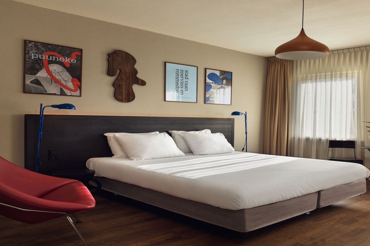 A modern hotel room with a large bed, a red chair, a small table with a plant, and artwork on the wall.