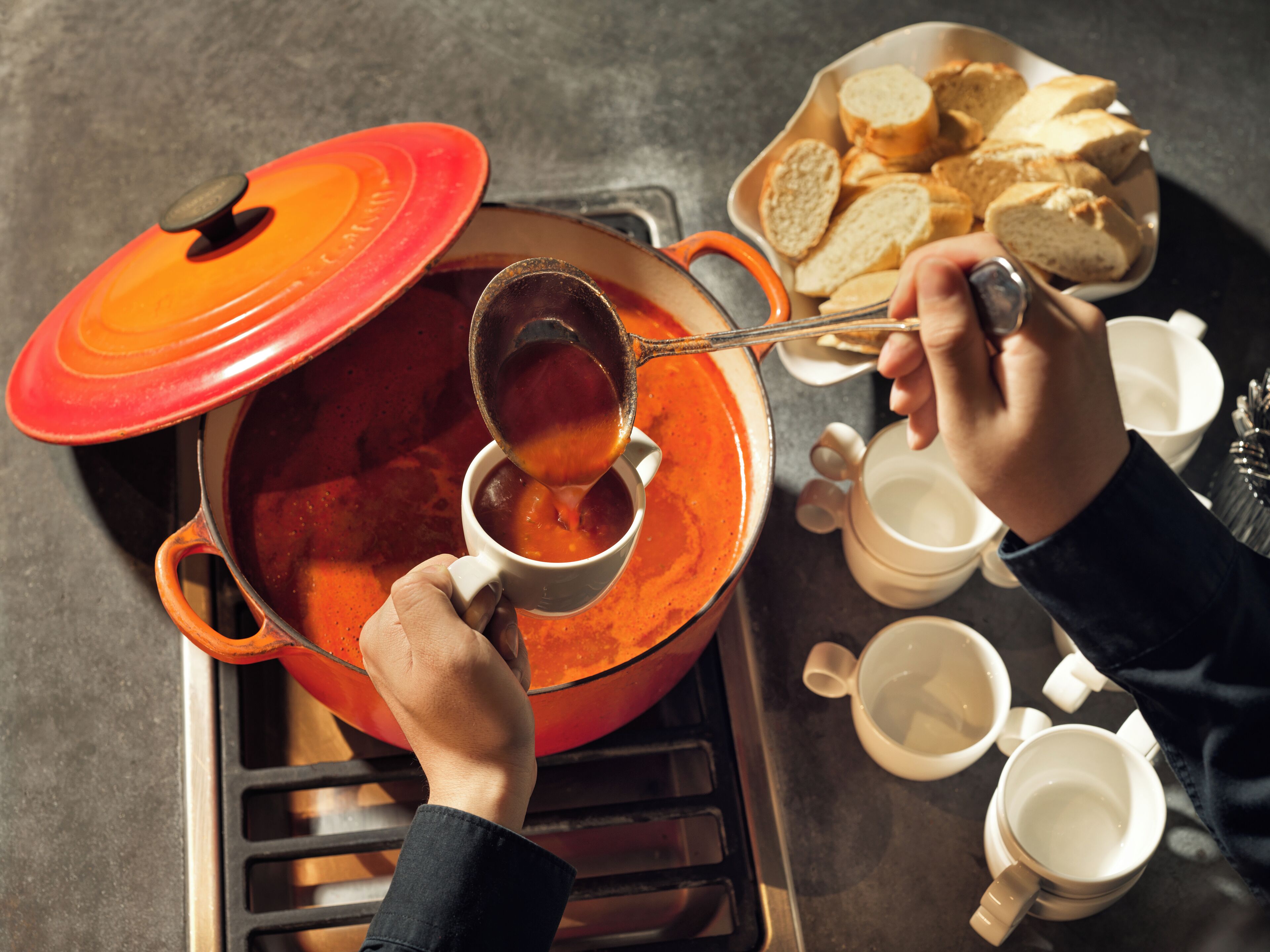 A person cooking soup in a red pot on a stove, with bread and cups nearby.