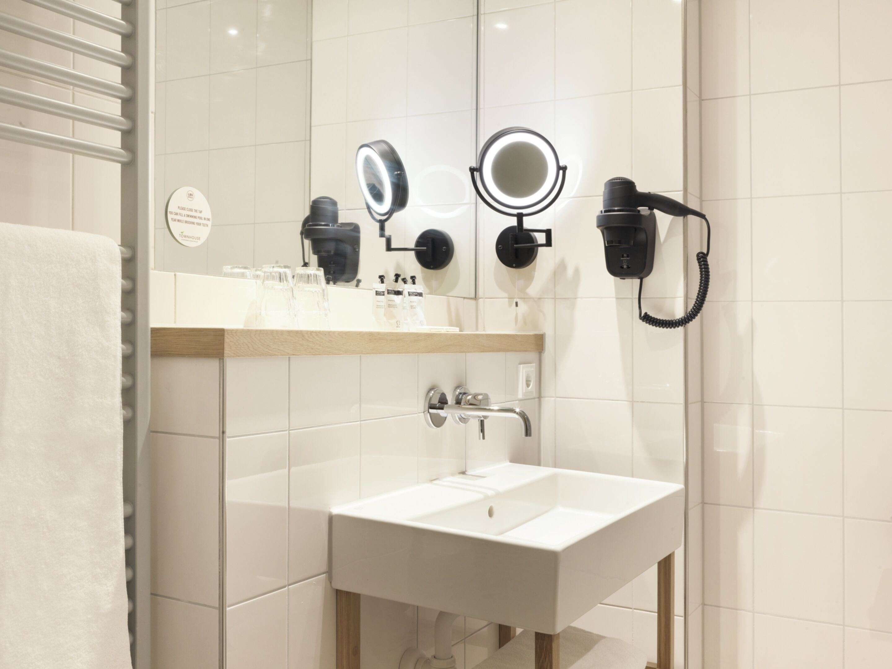 A modern bathroom with a white sink, two wall-mounted hair dryers, and a mirror with a light.
