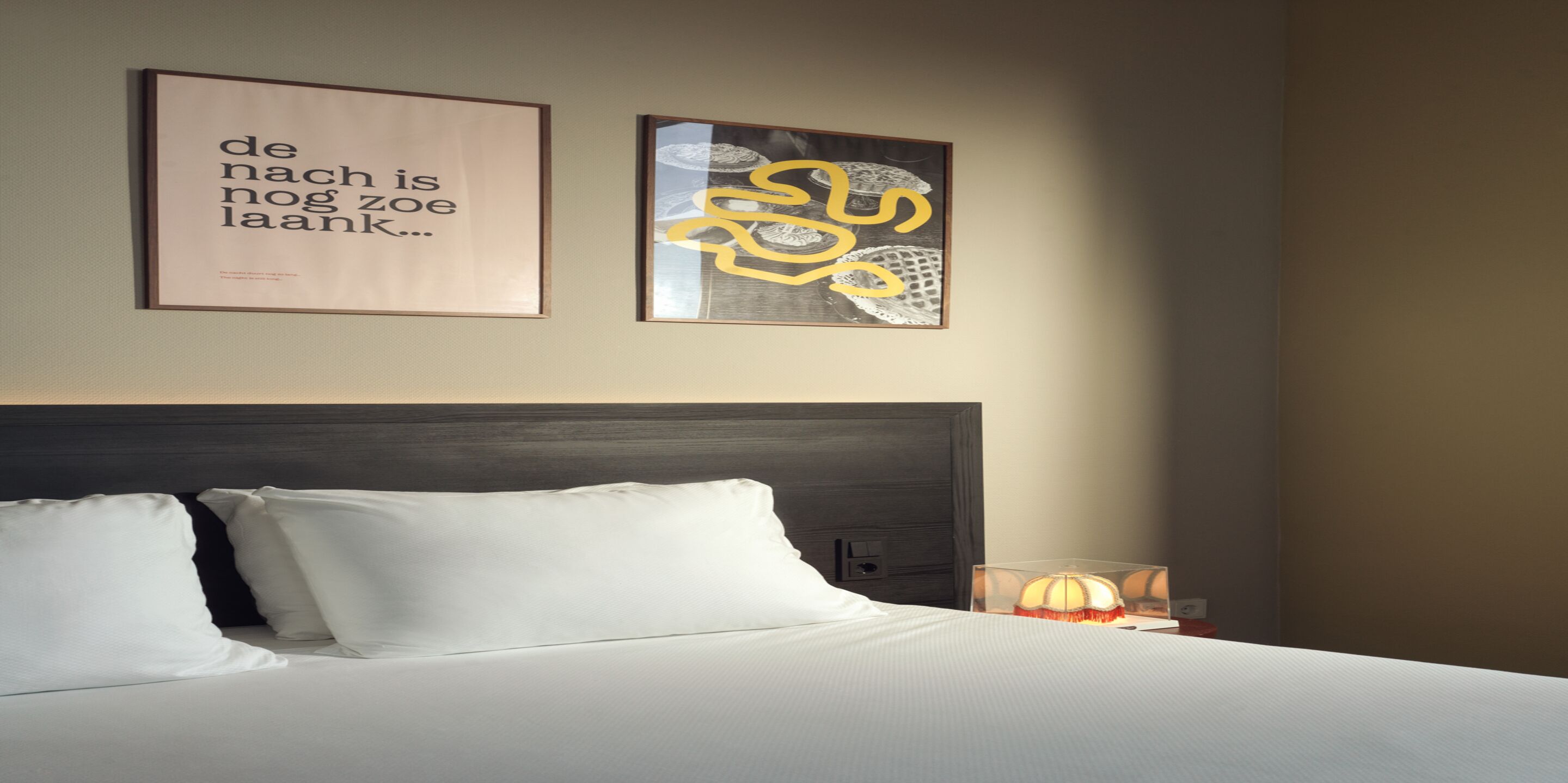 Una cama bien tendida con sábanas y almohadas blancas, un cabecero oscuro y dos cuadros enmarcados en la pared.