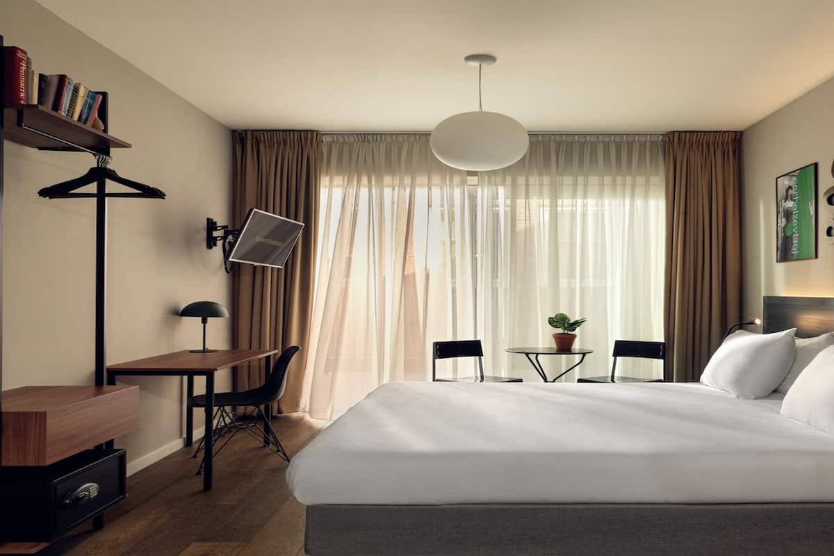 A hotel room with a large bed, a desk with a chair, a bookshelf, a TV, and a window with curtains.