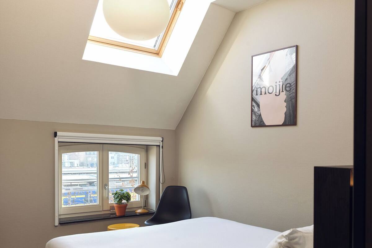 A bedroom with a skylight, a bed, a chair, a desk with a lamp, and a window with a view of buildings.