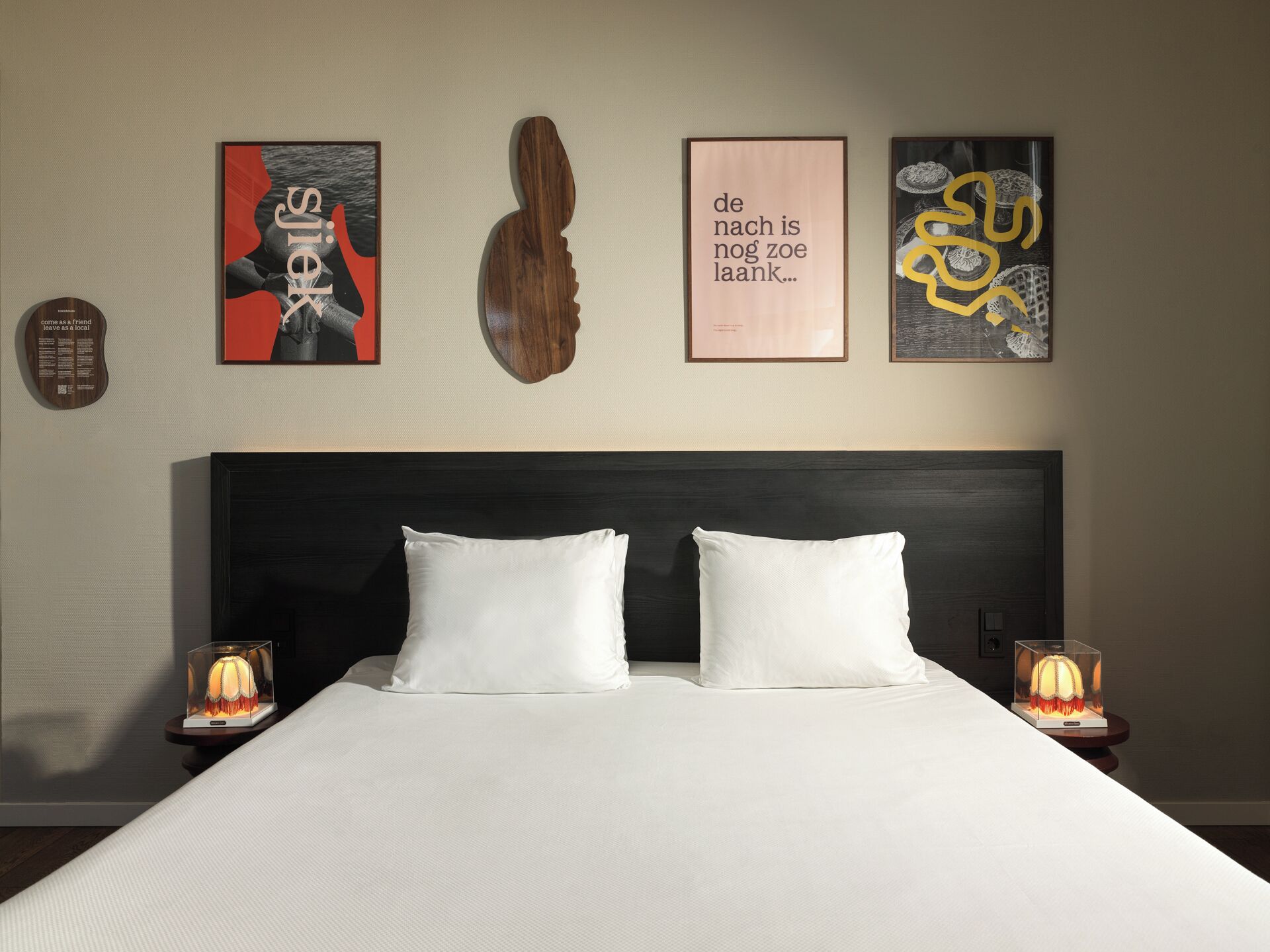 A hotel room with a large bed, two white pillows, a dark headboard, and three framed artworks on the wall.