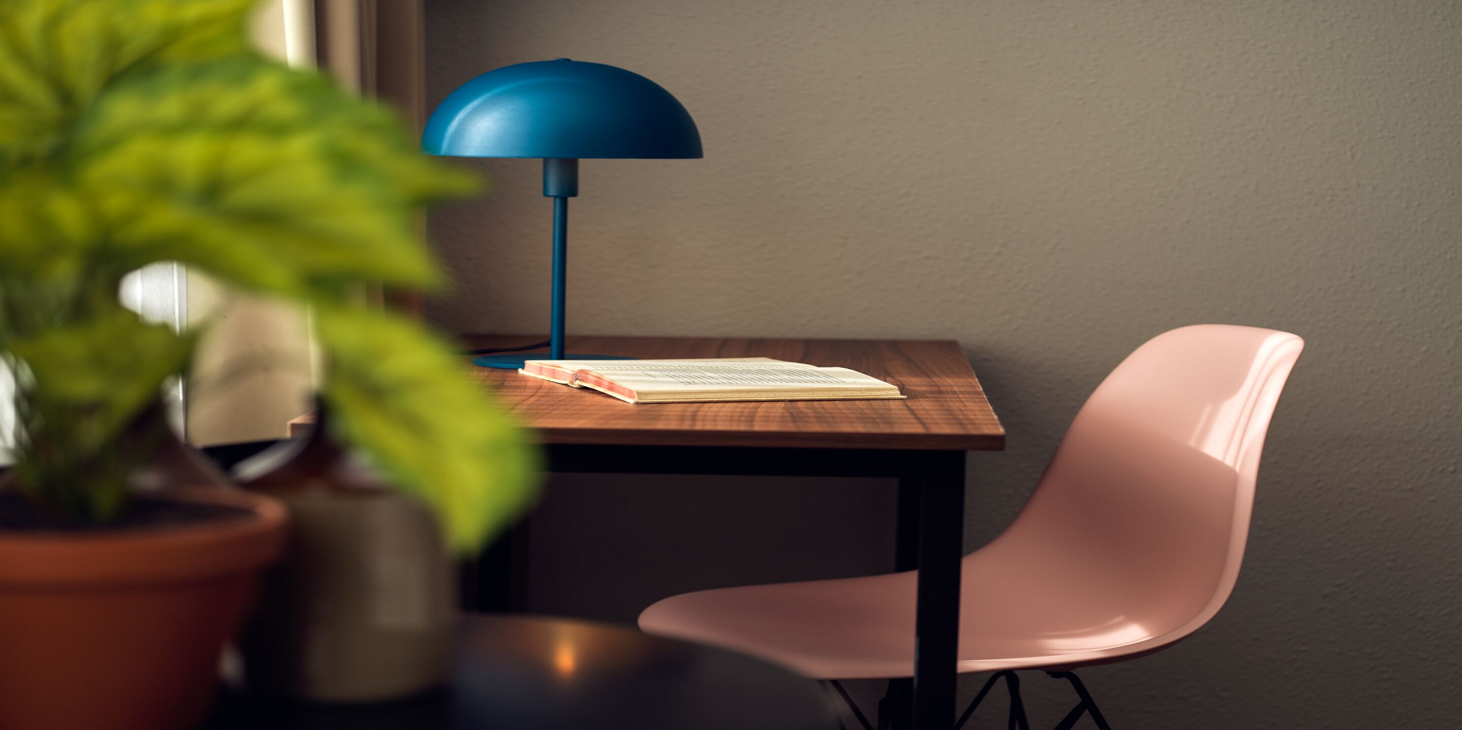 A wooden table with a blue lamp, a pink chair, and a book.