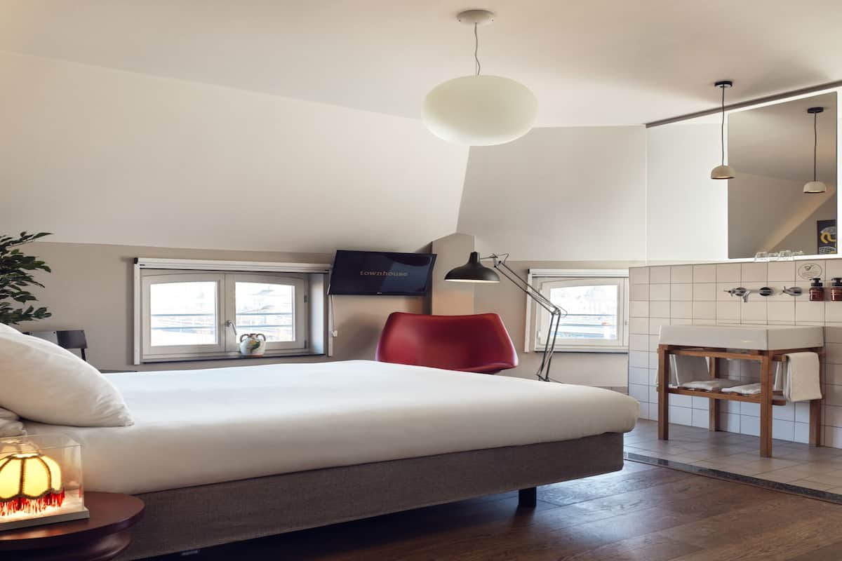 A modern bedroom with a large bed, bedside lamp, and a small table with a lamp. There is a red chair, a plant, and a window with a view.